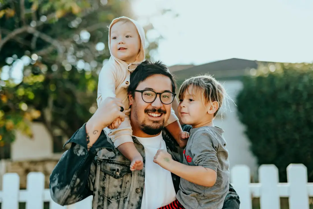 Pai sorrindo com os dois filhos no colo, no quintal de casa